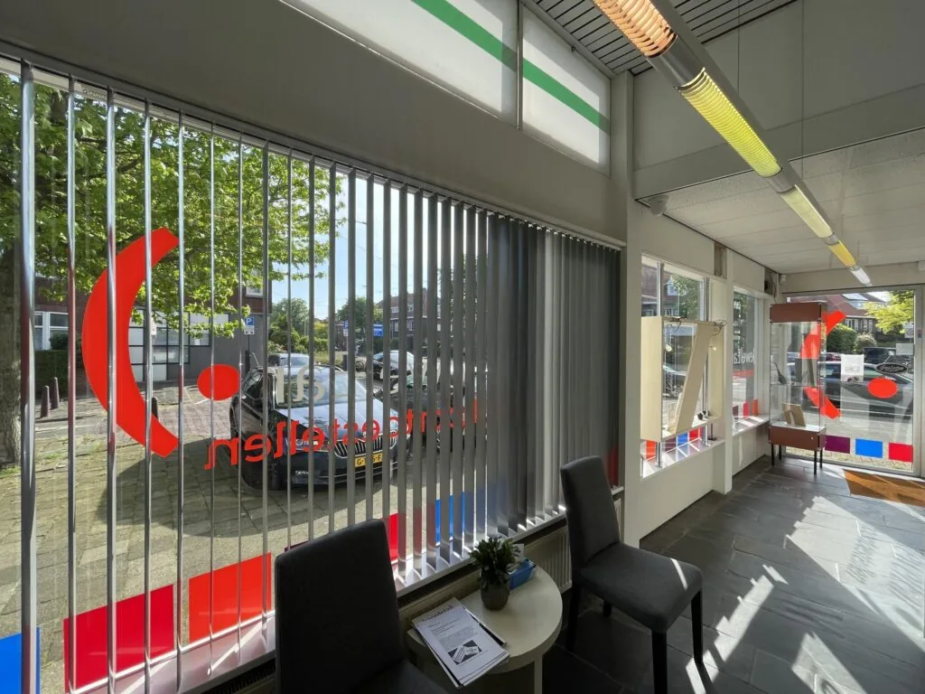 Winkelpand aan de Prinses Mariannelaan met grote ramen, verticale jaloezieën en kleurrijke raamstickers.